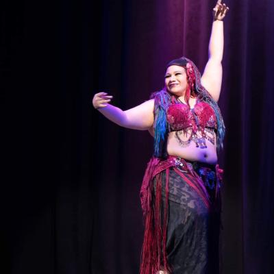 Female dancer on stage wearing dark red and black with mid-drift and arms exposed. Wearing blue and red yarn dreads. 