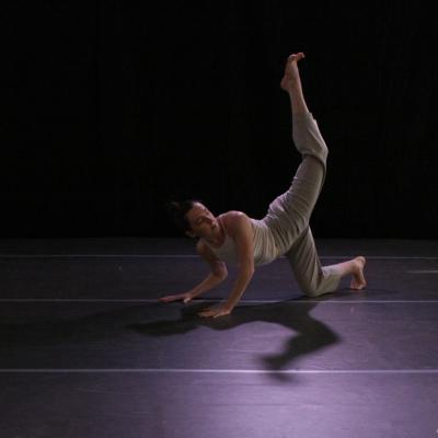 white woman balancing on her right knee with hands on the floor and left leg extended behind her in the air; wearing a white tank top and gray pants on a dark stage with a dim purple spotlight