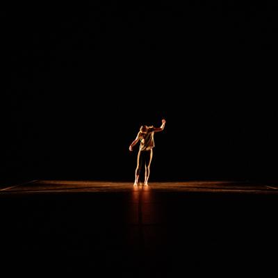 A dancer (Zoey November) stands alone on a dark stage, her body lit by a dim yellow light. One arm is in the air and her head is turned away in an expression of anguish
