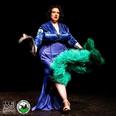brunette Caucasian performer in blue dress walking forward swinging a silver glove and holding a green ostrich feather boa