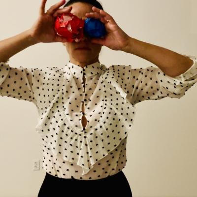 a woman in a white top with black polka dots and ruffles stands facing front. She holds one blue ball and one red ball in front of her eyes